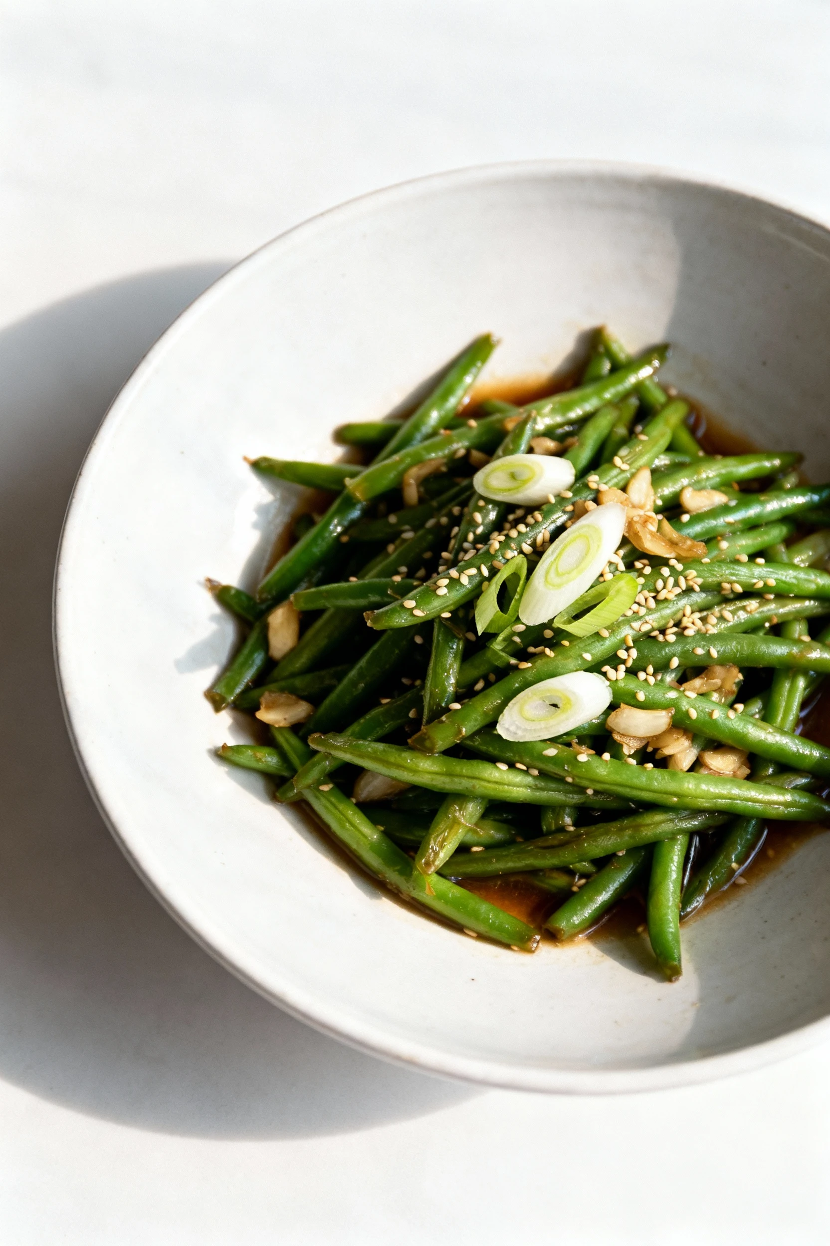 Overhead shot of freshly plated Asian garlic green bean stir-fry in a wide white ceramic bowl, garnished with sliced sca